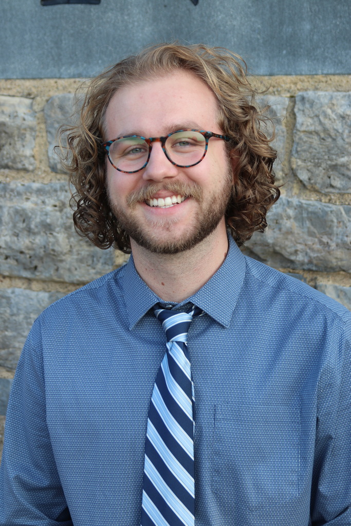 Man standing by stone wall in blue shirt and striped tie smilling.