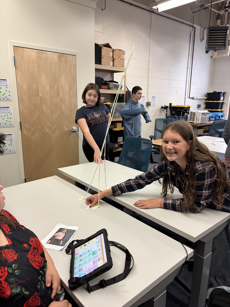 Two students standing and one seated around a table building a staw tower.