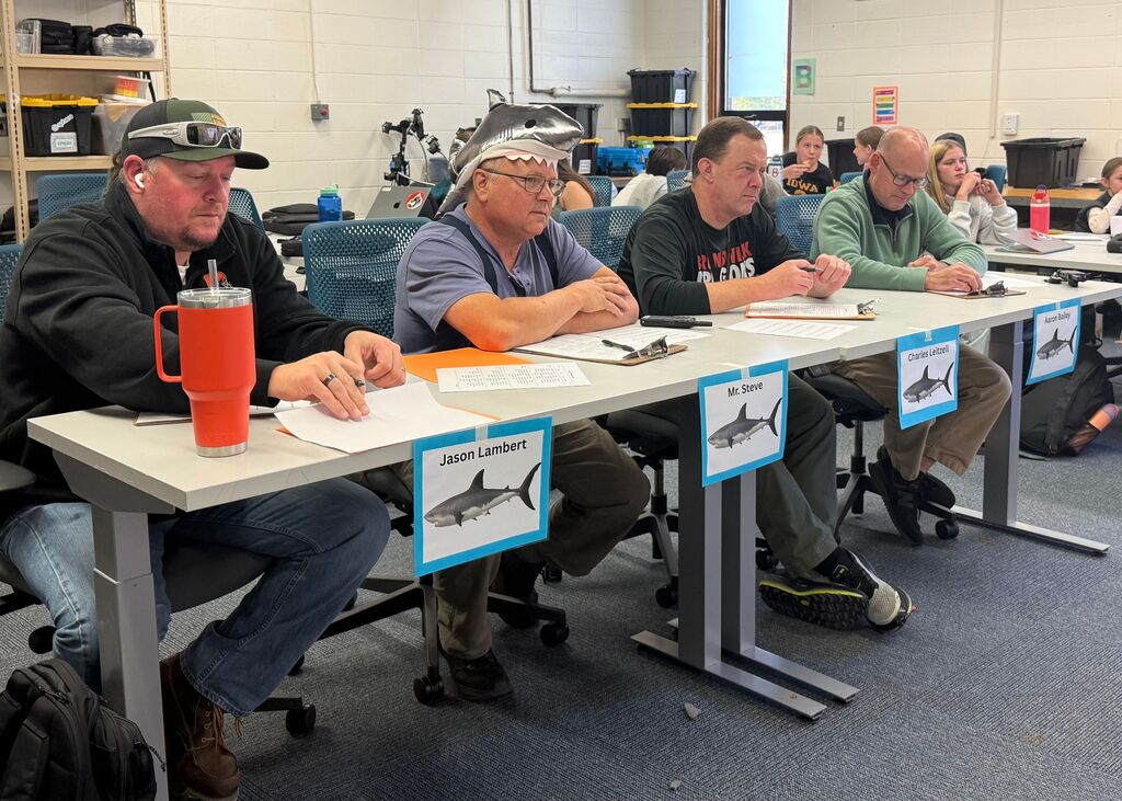 Table of 4 adults sitting at a long table, posing as "Sharks" for Shark Tank/