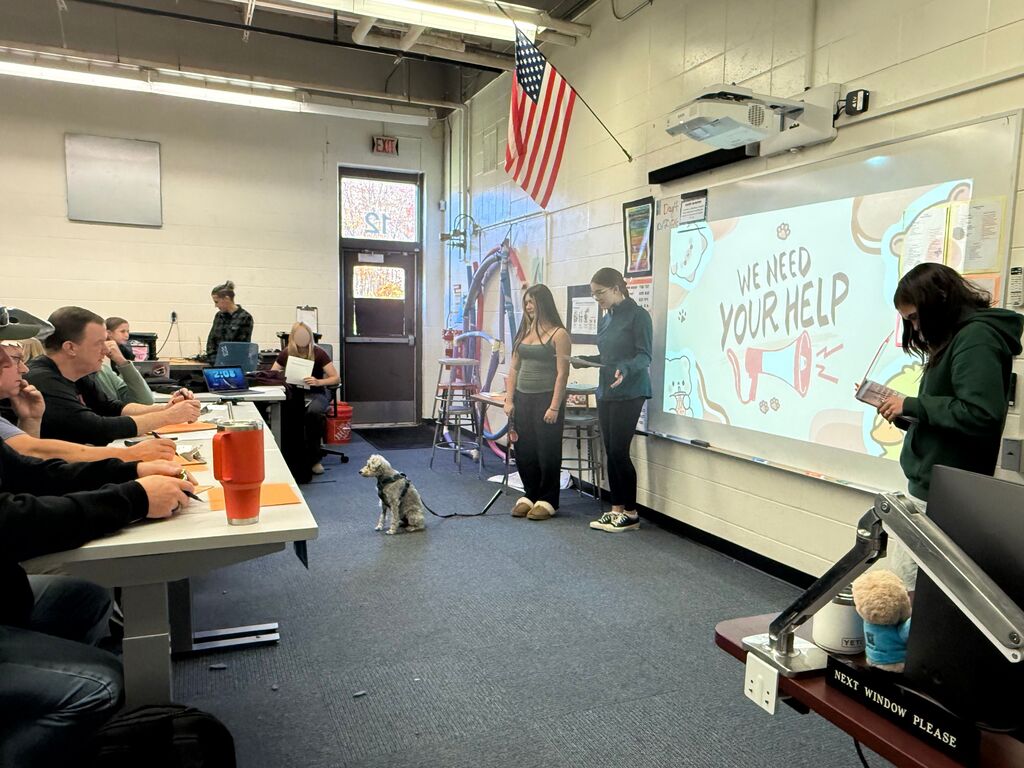 Three students giving a presentation with a dog in the front of the classroom.