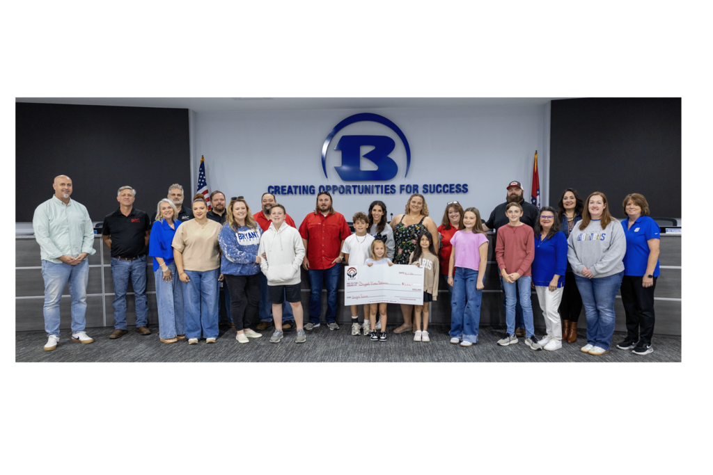 Group of people in casual attire, two holding a check, standing in front of a company logo and flag.