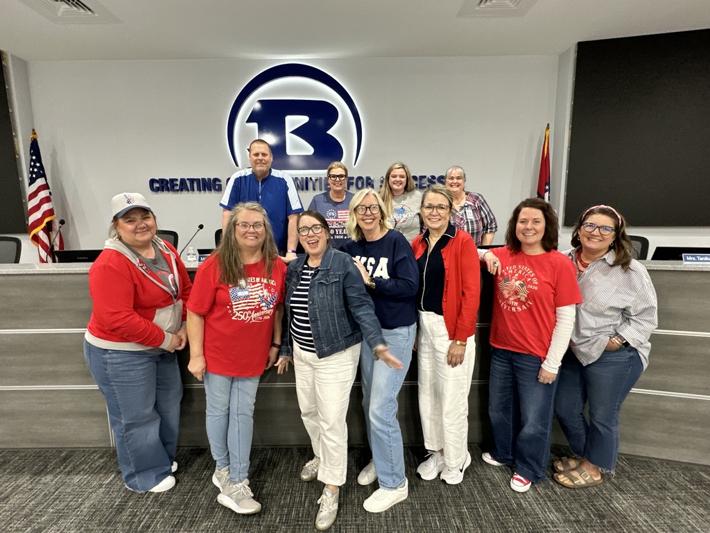 Group of people wearing red shirts, standing in a conference room. Two flags are visible in the background.