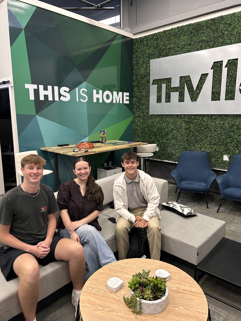 Three people sit on a couch, two women and one man, with a table, plant, and chairs in a room. A sign behind them reads "This is Home."