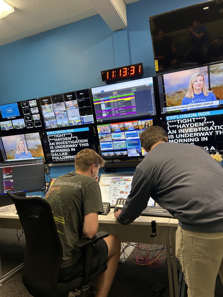Two individuals working in a control room, facing screens with news and other content.