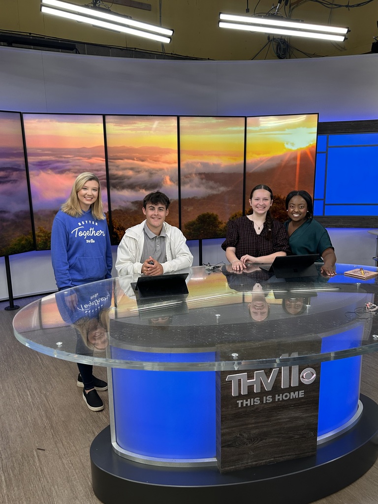 Four individuals stand behind a glass news desk. Behind them, a large screen displays a sunset.