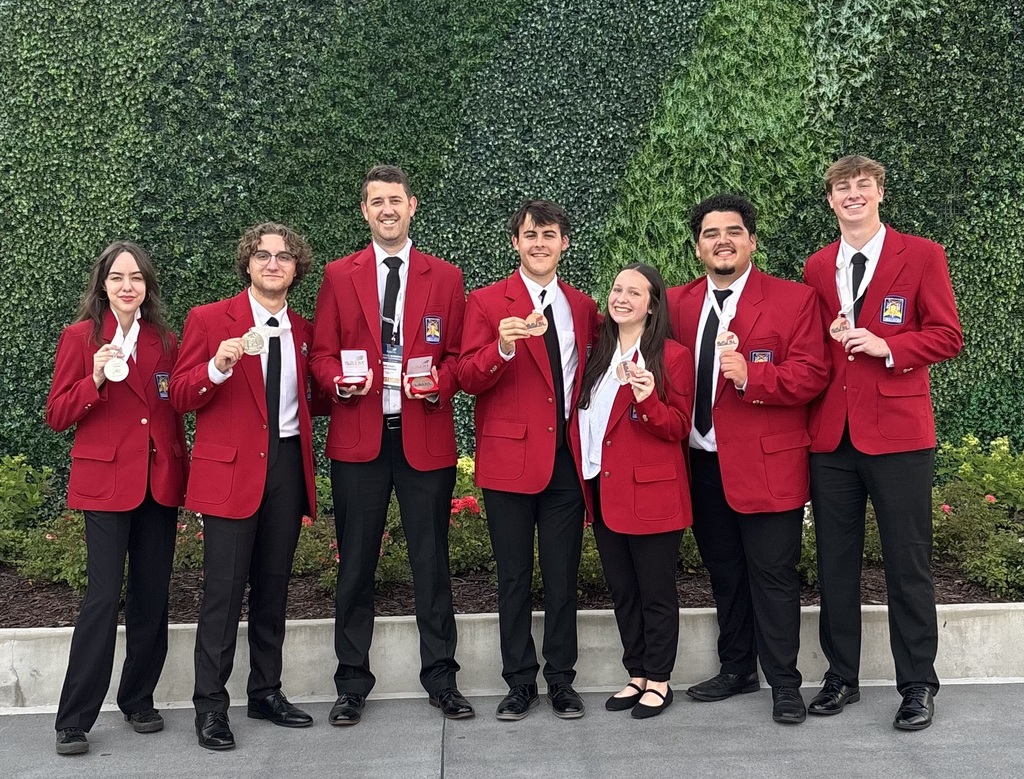 Eight people, wearing red blazers and black bottoms, pose together for a photo in front of a green wall.