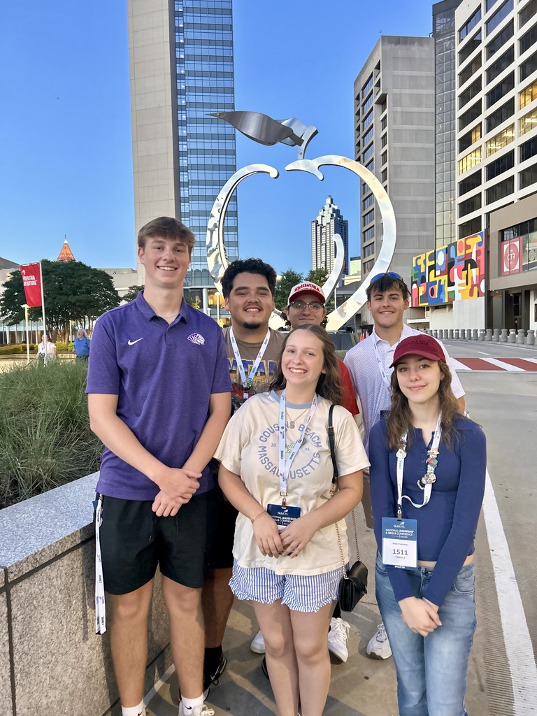 Six young adults wearing ID badges pose in front of a large silver apple sculpture and buildings.