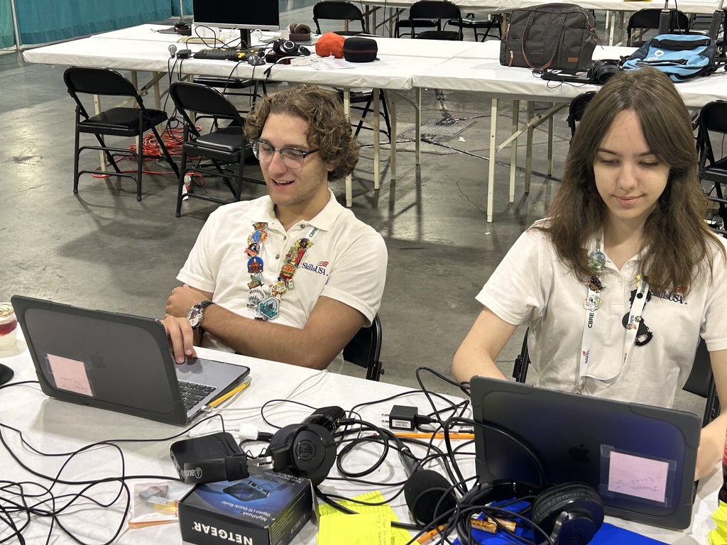 Two people sit at a table with laptops and headphones in front of them. Behind them are tables and chairs.