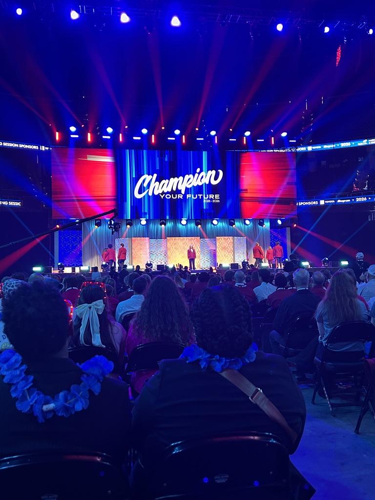 Audience seated in chairs at a stage, watching an event with blue and red lighting and a large screen displaying "Champion".
