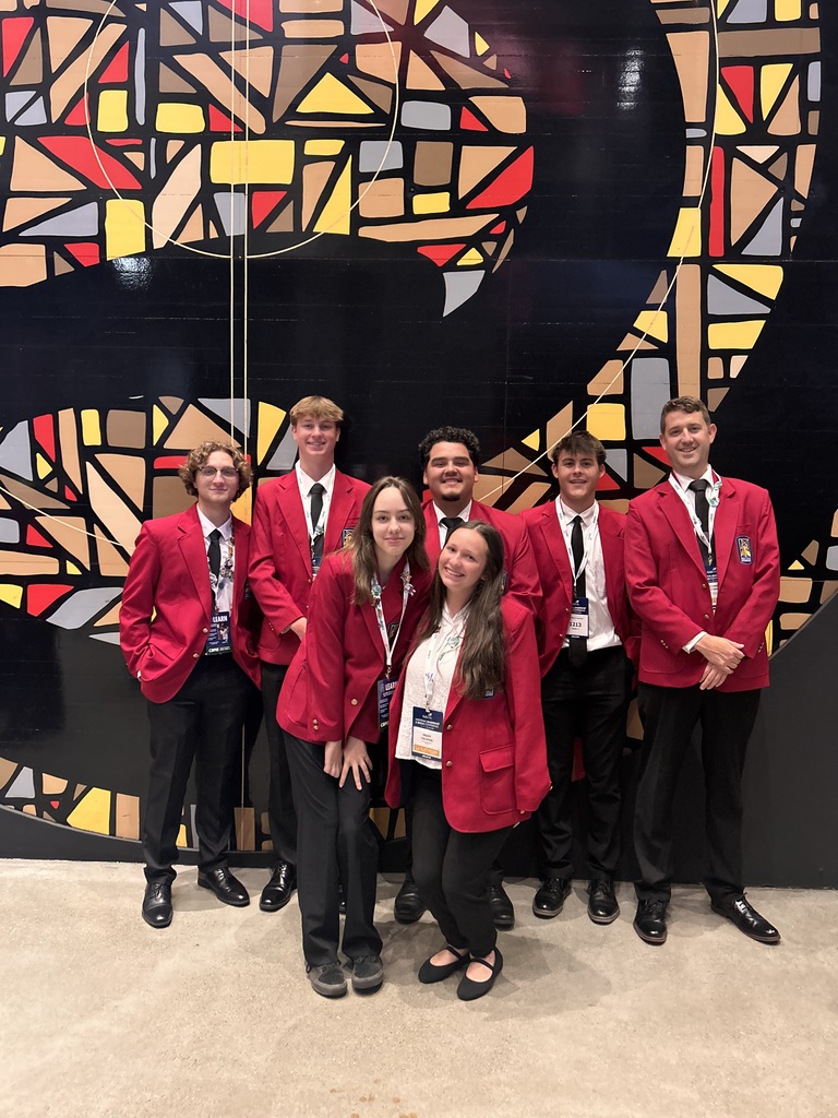 Seven people in red blazers and black pants pose for a photo in front of a colorful geometric wall.