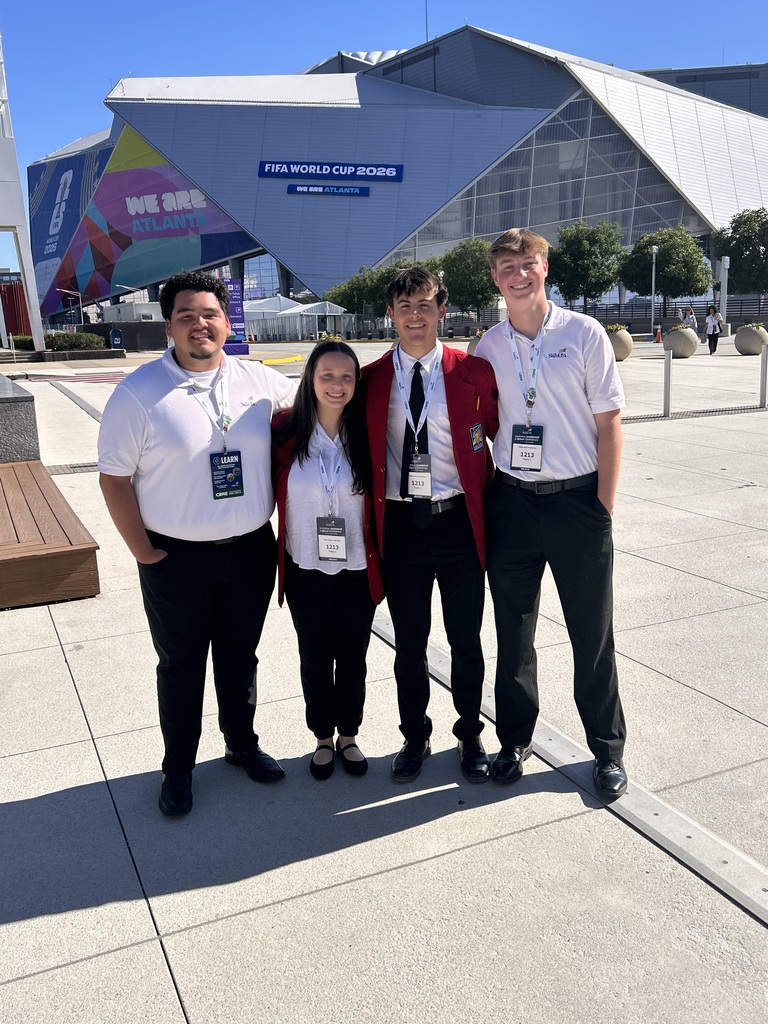 Four individuals in matching shirts and ties standing in front of a building with a sign.