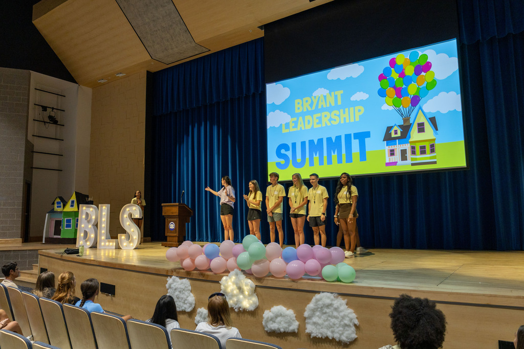 Five girls in yellow shirts stand on stage, presenting at an event. Behind them, a large screen displays "Brilliant Leadership Summit".
