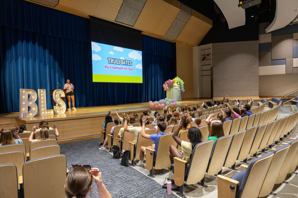 Audience seated in rows at an event. A person stands on stage by large letters. A projector screen displays text.