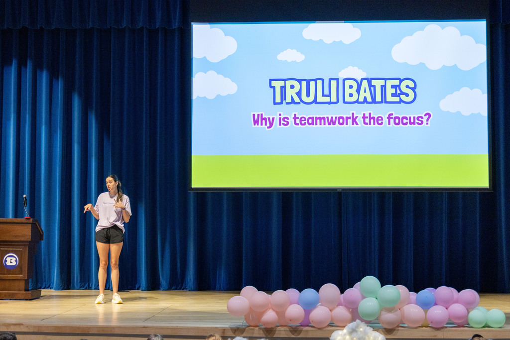 A woman in a white shirt and shorts speaks at a podium on a stage with blue curtains. A screen behind her displays "TRULI BATES Why is teamwork the focus?"