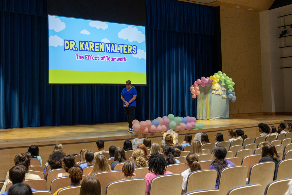 A person stands on a stage with balloons and a projector screen. The screen reads "Dr. Karen Walters." An audience sits in chairs.