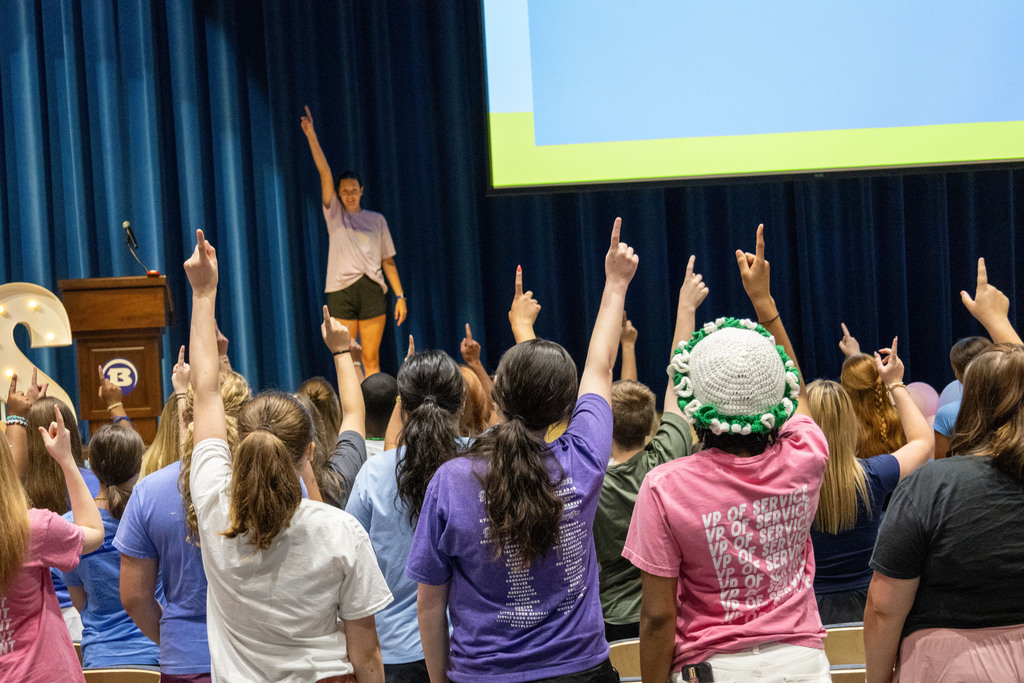 People in a room raising hands, one person standing in front with hands raised, blue curtain and screen behind.