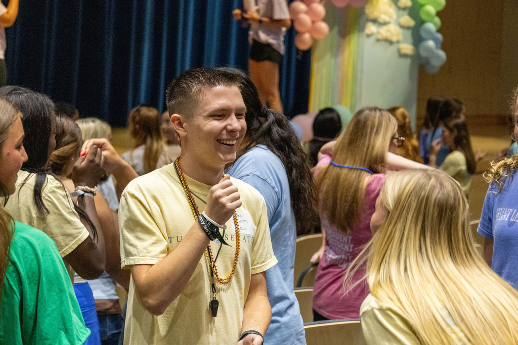Several people, some in light-colored T-shirts, stand indoors. A man smiles in front of a group.