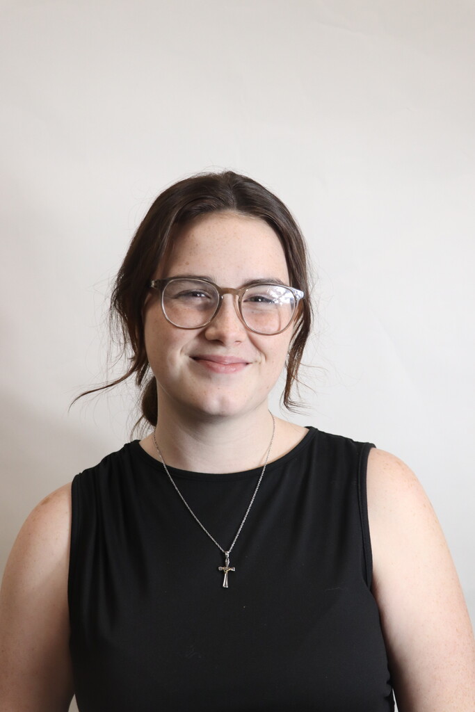 A woman with glasses and a necklace with a cross pendant smiles at the camera. She wears a black sleeveless top against a white background.