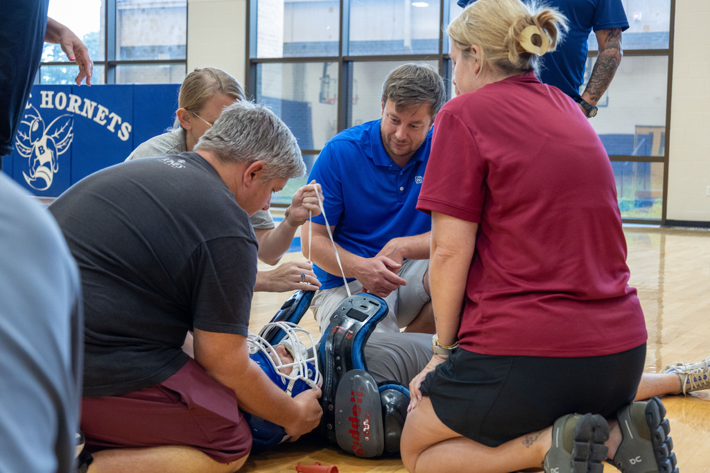 Several people in a gymnasium. One person kneels on the floor adjusting the leg protection of another.