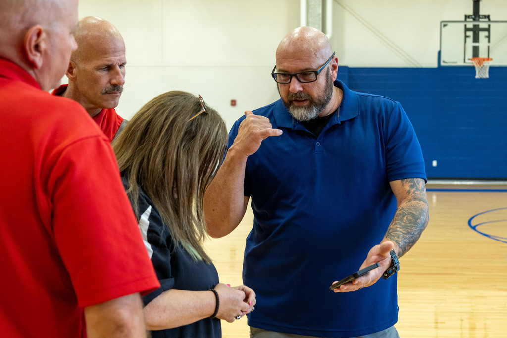 A group of people standing in a basketball court, one man showing something on a phone.