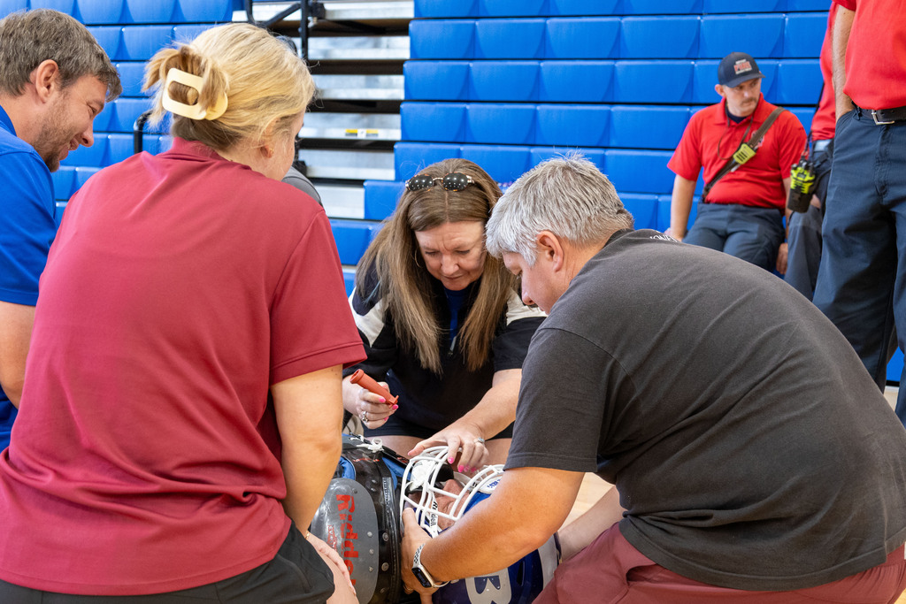 Six people in red, blue, and black shirts are in a gym. Three are seated; one holds a football helmet.