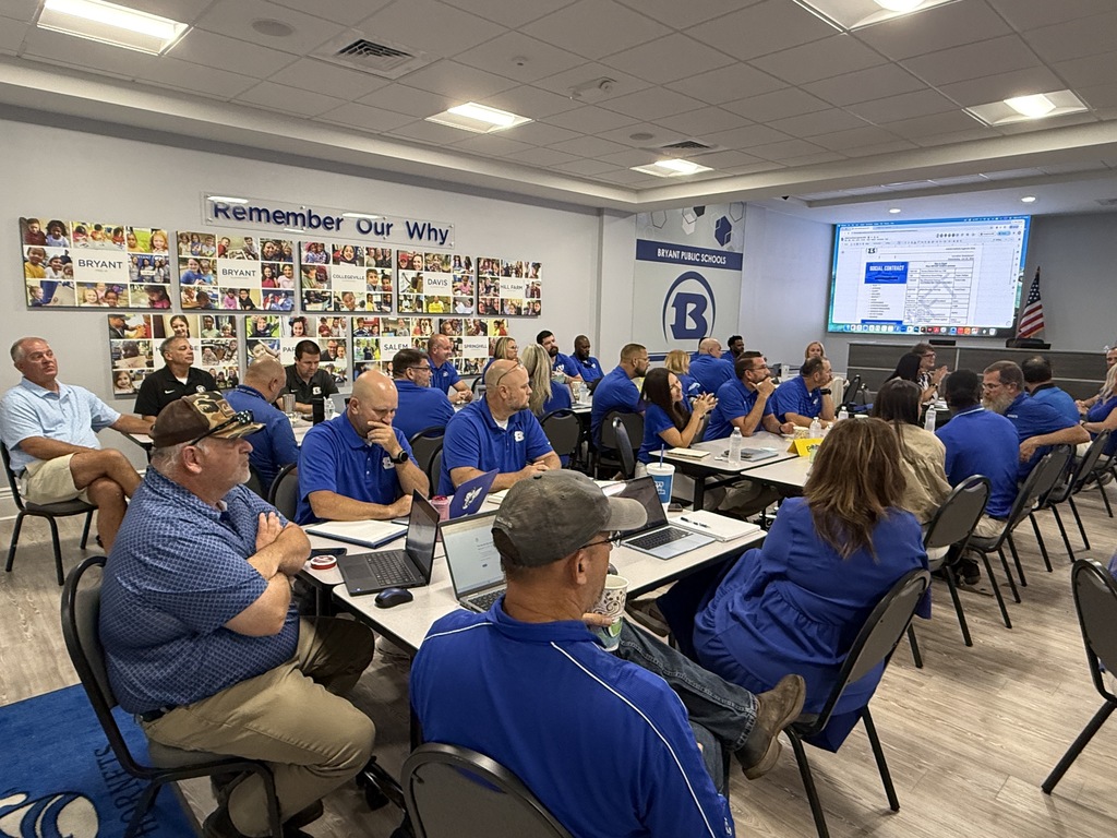 A group of people in blue uniforms sitting at desks with laptops, a projector screen in the room.
