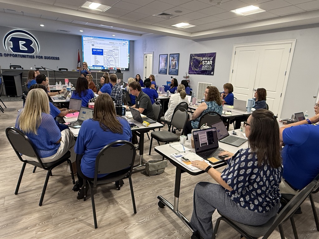 People in blue shirts sit around tables with laptops in a room with a projector screen.