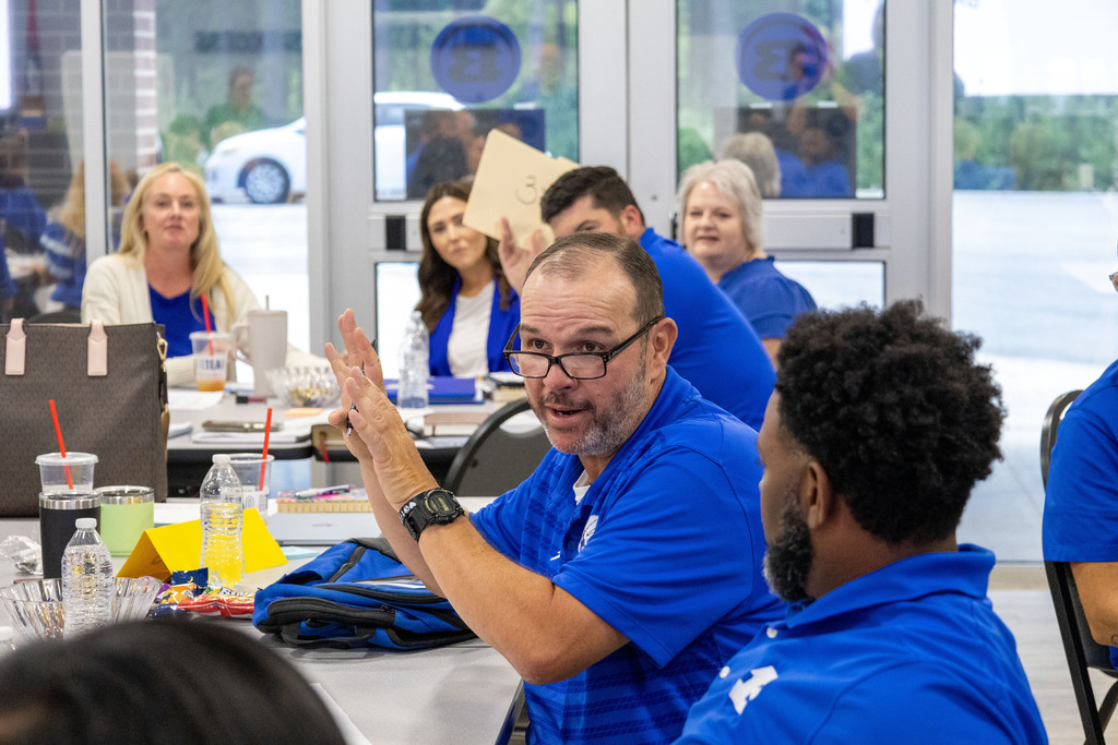 Group of people in blue shirts seated at a table, engaging in conversation and smiling.