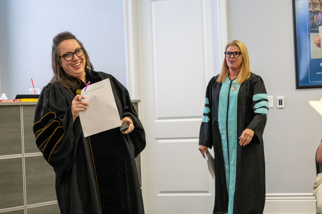 Two women in academic robes, one holding papers, in a room with a desk and framed picture.