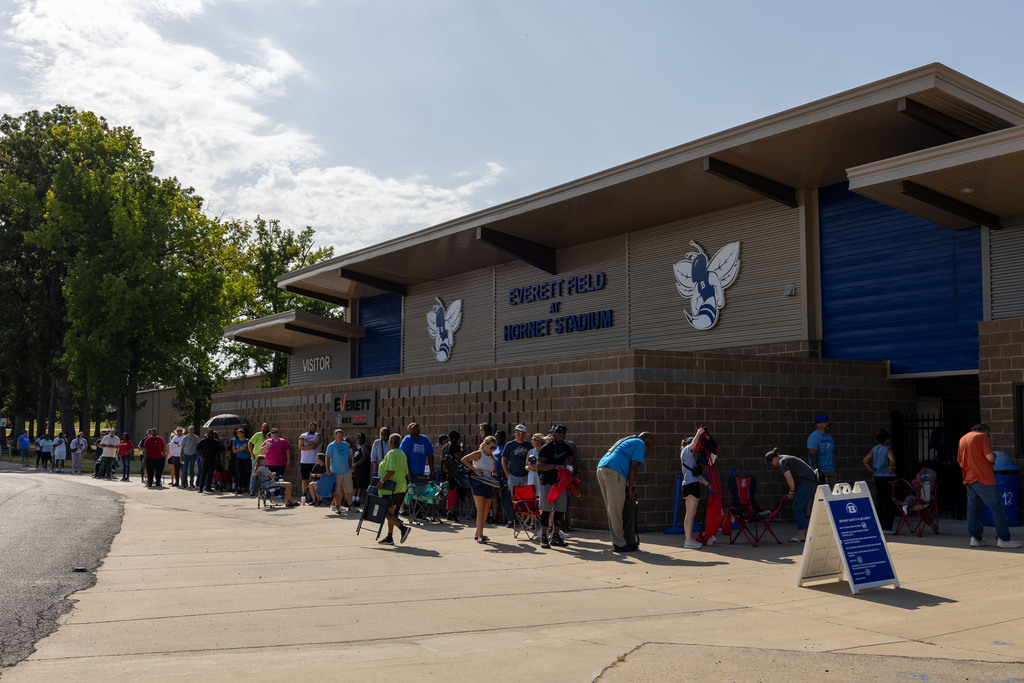 People wait in line outside a building with a blue roof and emblems on the side.