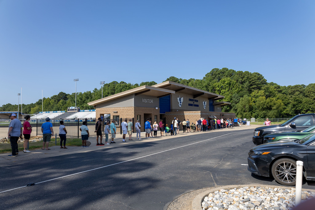 People wait in line at a building. Trees and a stadium are visible in the background. Cars are parked nearby.