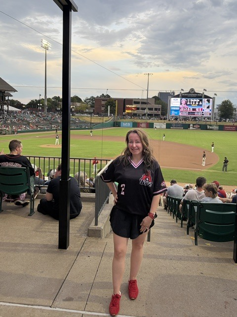 A woman in a black jersey smiles and stands at a baseball game, with empty bleachers and a large screen behind her.