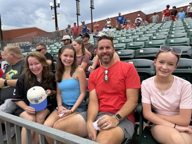 Four people seated in stadium bleachers. Three women, one in a black shirt, and a man in a red shirt.