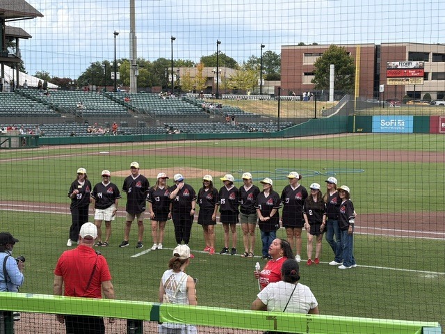 A group of people, some in hats, stands on a baseball field with spectators watching from behind a barrier.