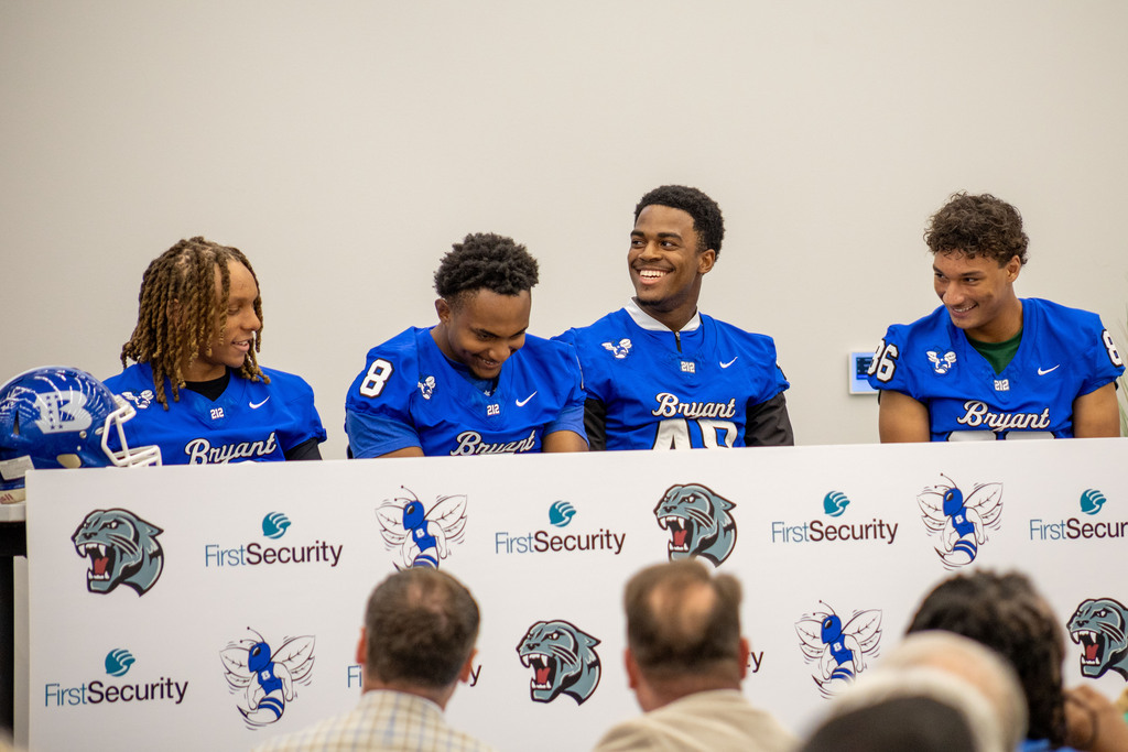 Four athletes sit behind a table with a panel of logos. The athletes wear blue jerseys.