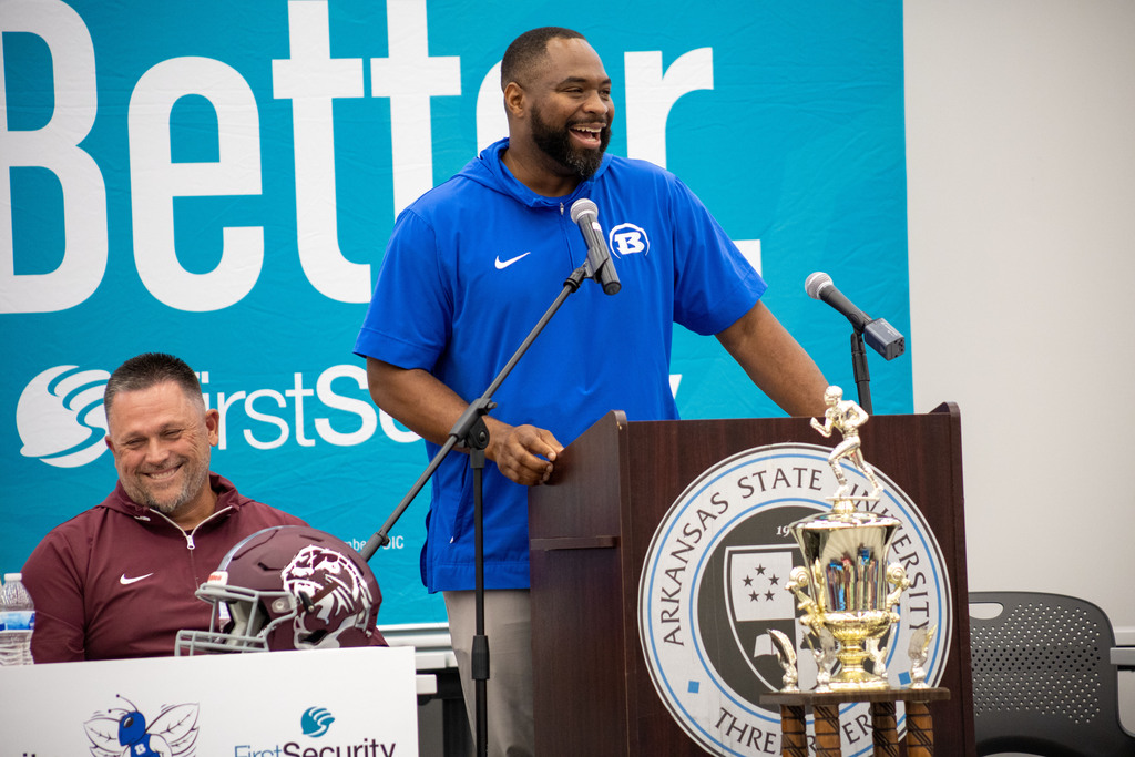 Two men stand at a podium with a trophy. One man in a blue shirt holds a microphone, while the other sits behind a table.