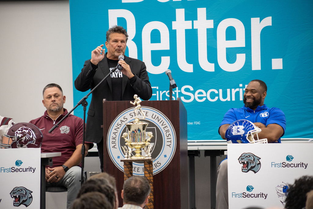 A man speaks at a podium with a trophy in front of him. Two men sit behind him, one with a football helmet.
