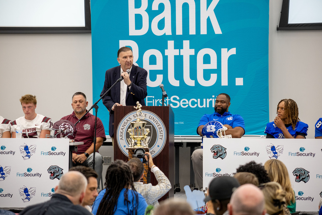A man speaks at a podium with a Bank Better banner behind him. Several people sit at tables.
