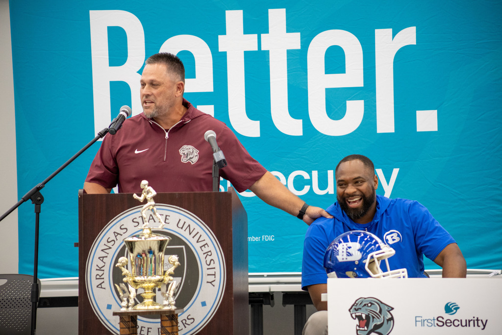 Two men at a podium, one seated and one standing, with a microphone and trophy, banner reads "Better".