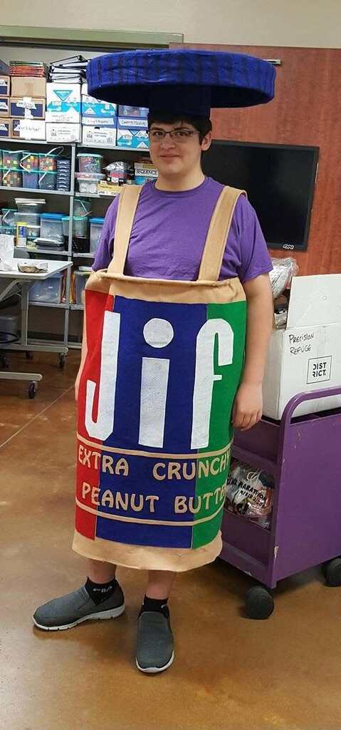Person dressed as a Jif peanut butter container with a hat, standing in a room with shelves and a television.
