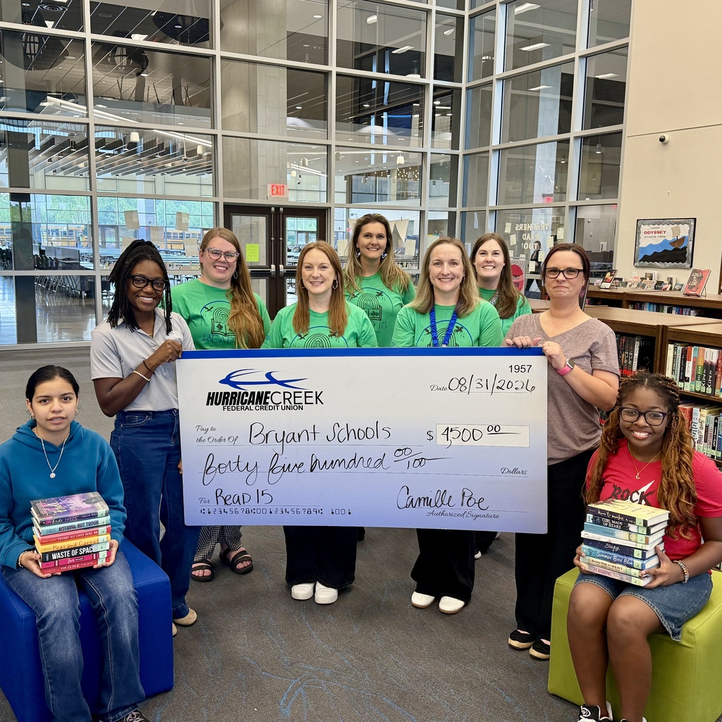 A group of students holding a check and books in a library with bookshelves and large windows.