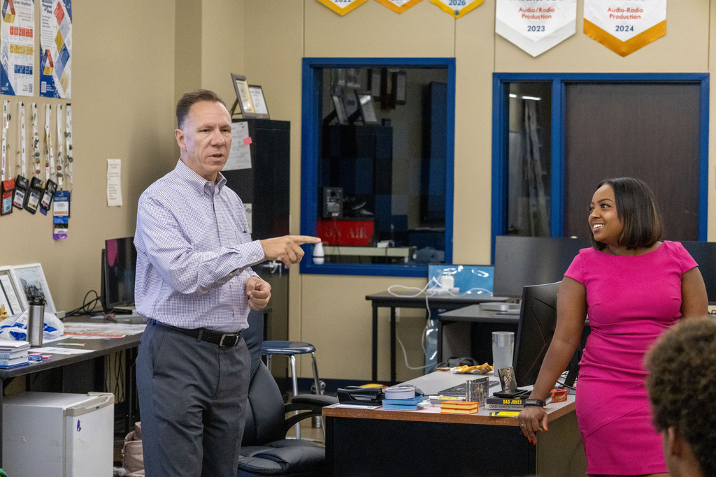 A man points at a woman standing at a desk in a room with desks, chairs, and a whiteboard.