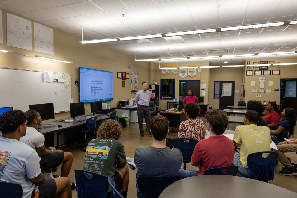 A classroom with a teacher standing, addressing students seated in a circle around a table. A large monitor is on the wall.