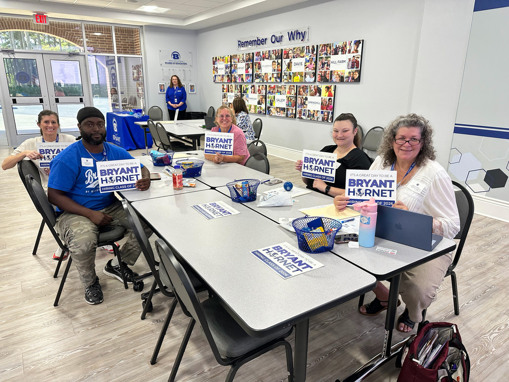 People seated around a table hold signs reading "Bryant Helmet". One person wears a blue shirt. Behind, a wall displays photos.