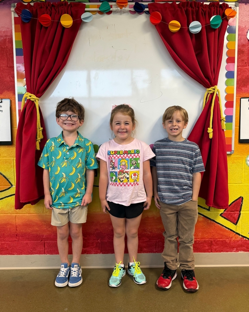 Three children stand in front of a decorated wall with red curtains and a colorful backdrop.