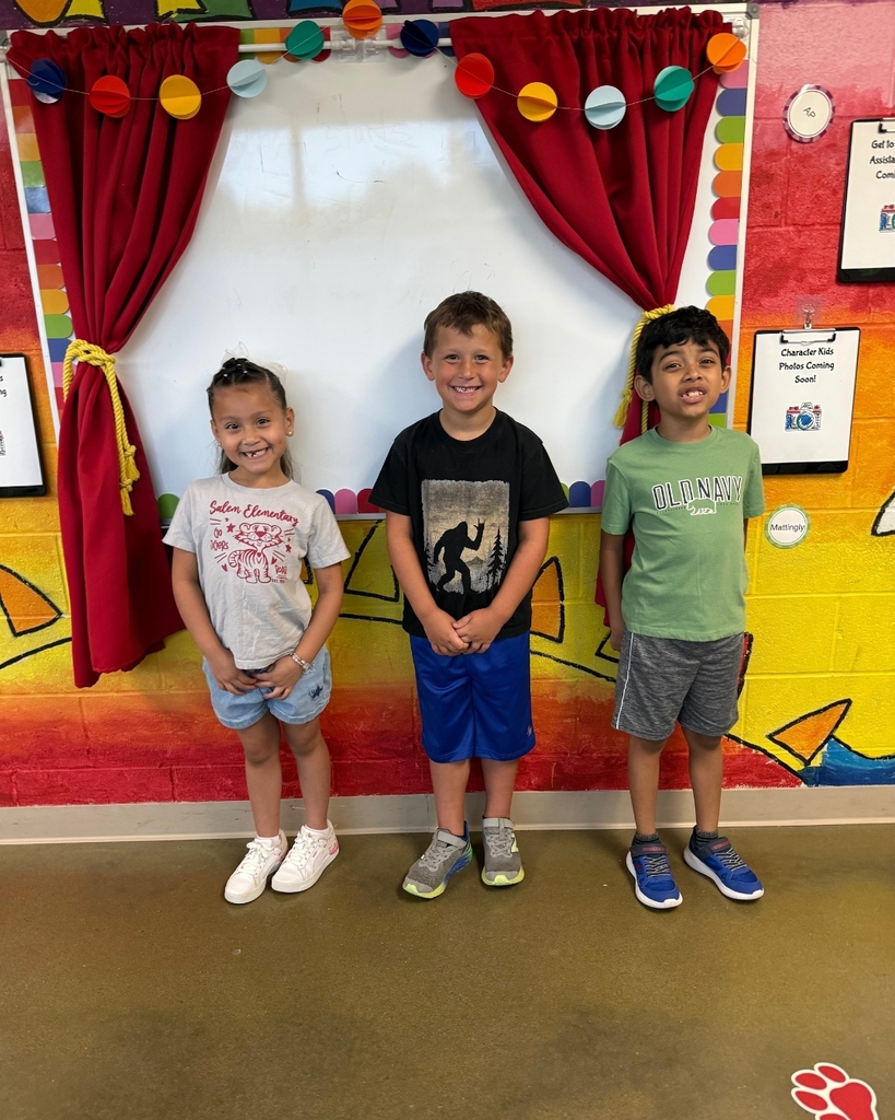 Three children standing in front of a colorful wall with shapes and pictures. A white backdrop with red curtains is behind them.