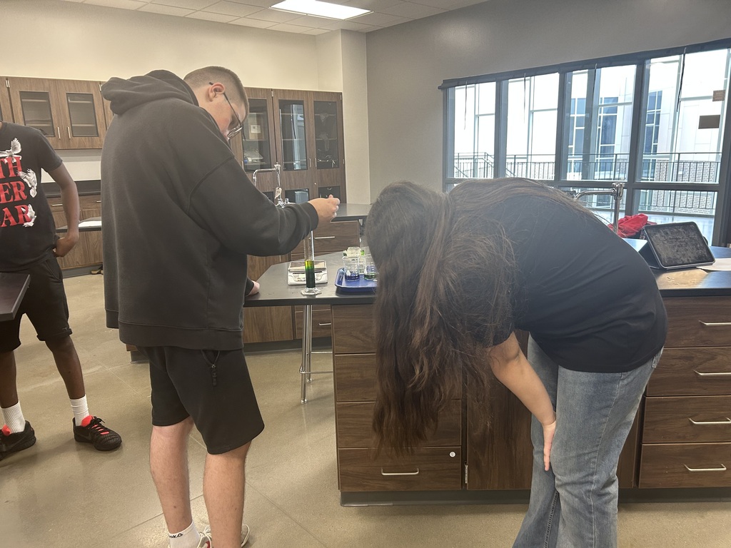 Two students working at a lab table with cabinets, one bending over while the other stands.