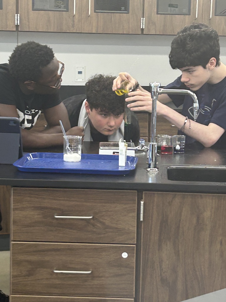 Three students are seated at a counter in a lab. One adds liquid to a beaker.
