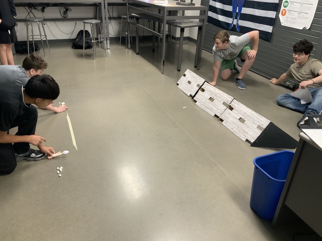 Four students on a floor, working on an activity, holding paper, and one person kneeling. Background includes a table, trash bin, and a bulletin board.