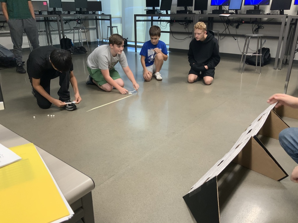 Four young individuals work on a project in a room with a white floor. One kneels, and another holds a long object.